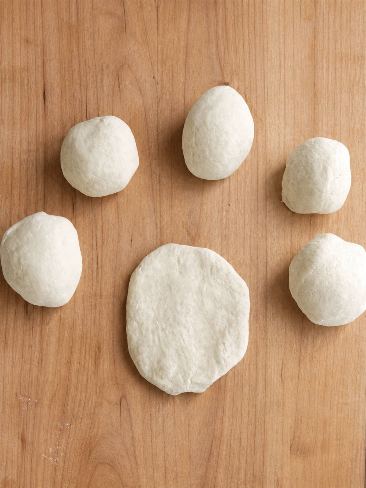 Flour tortilla dough balls and one flattened dough disc on a wooden board.