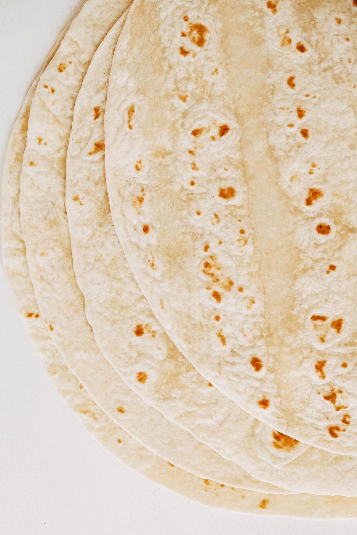 Several overlapping flour tortillas are arranged in a fan shape on a white surface, showing lightly toasted brown spots on their soft, thin surfaces.