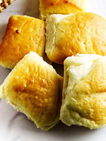 A basket filled with fluffy, golden-brown dinner rolls sits on a white surface.