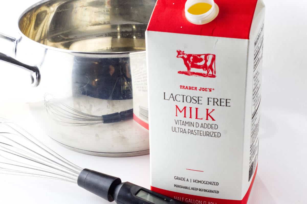 A carton of lactose free milk next to a stainless steel pot, a digital thermometer, and a whisk.