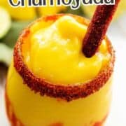 A glass of bright yellow Mangonada smoothie with red chamoy sauce swirls, topped with a tamarind candy stick and chili-lime seasoning on the rim, with another chamoyada, lime slices, and a green napkin in the background.