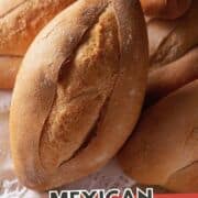 Four freshly baked Mexican bolillo rolls with golden, crispy crusts are stacked on a wooden surface. A red banner at the bottom reads “Mexican Bolillos” with a black website tag below.