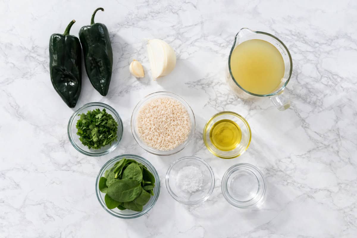 Overhead view of two poblano peppers, garlic, onion, chicken broth, rice, vegetable oil, cilantro, spinach, salt, and a small bowl of white powder arranged on a white marble surface.