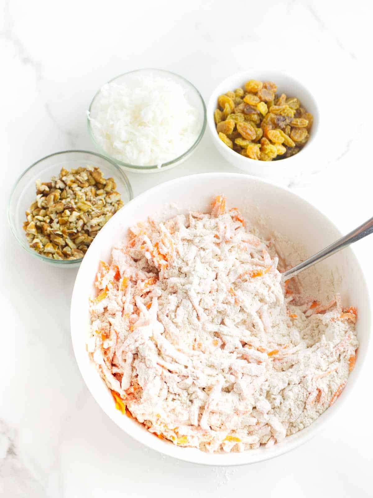 grated carrots coated with flour, and small bowls with raisins, chopped nuts, and shredded coconut nearby.