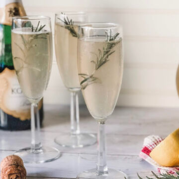 Three champagne flutes filled with a light bubbly drink, each garnished with a sprig of rosemary—perfect for pairing with quick and easy meals. A champagne bottle, corks, and a folded napkin rest in the background on a white surface.