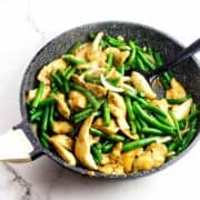 Bowl of stir-fried chicken, green beans, and onions with chopsticks; skillet and blue teapot on a white surface nearby.