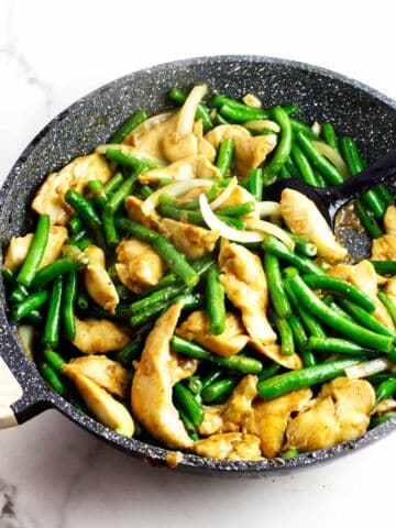 Bowl of stir-fried chicken, green beans, and onions with chopsticks; skillet and blue teapot on a white surface nearby.