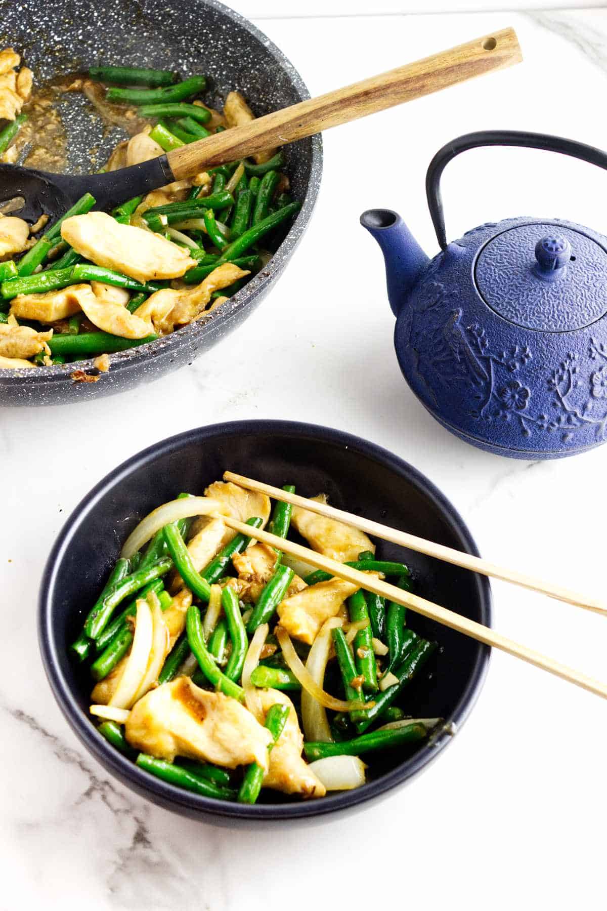 Bowl of Panda Express String Bean Chicken with green beans and onions, chopsticks, skillet, and blue teapot in background.