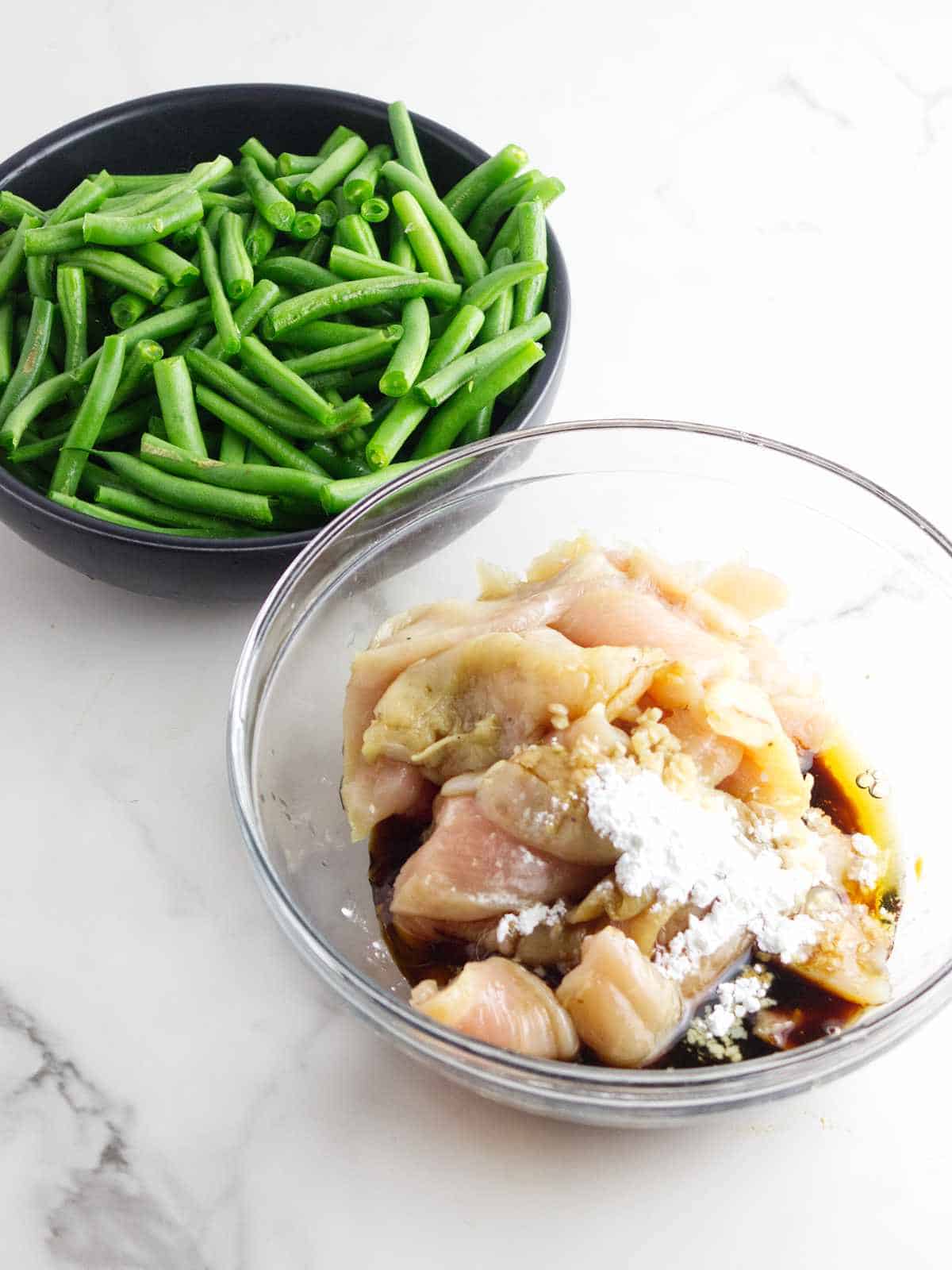 marinating chicken breast in a bowl, with trimmed green beans in a bowl nearby.
