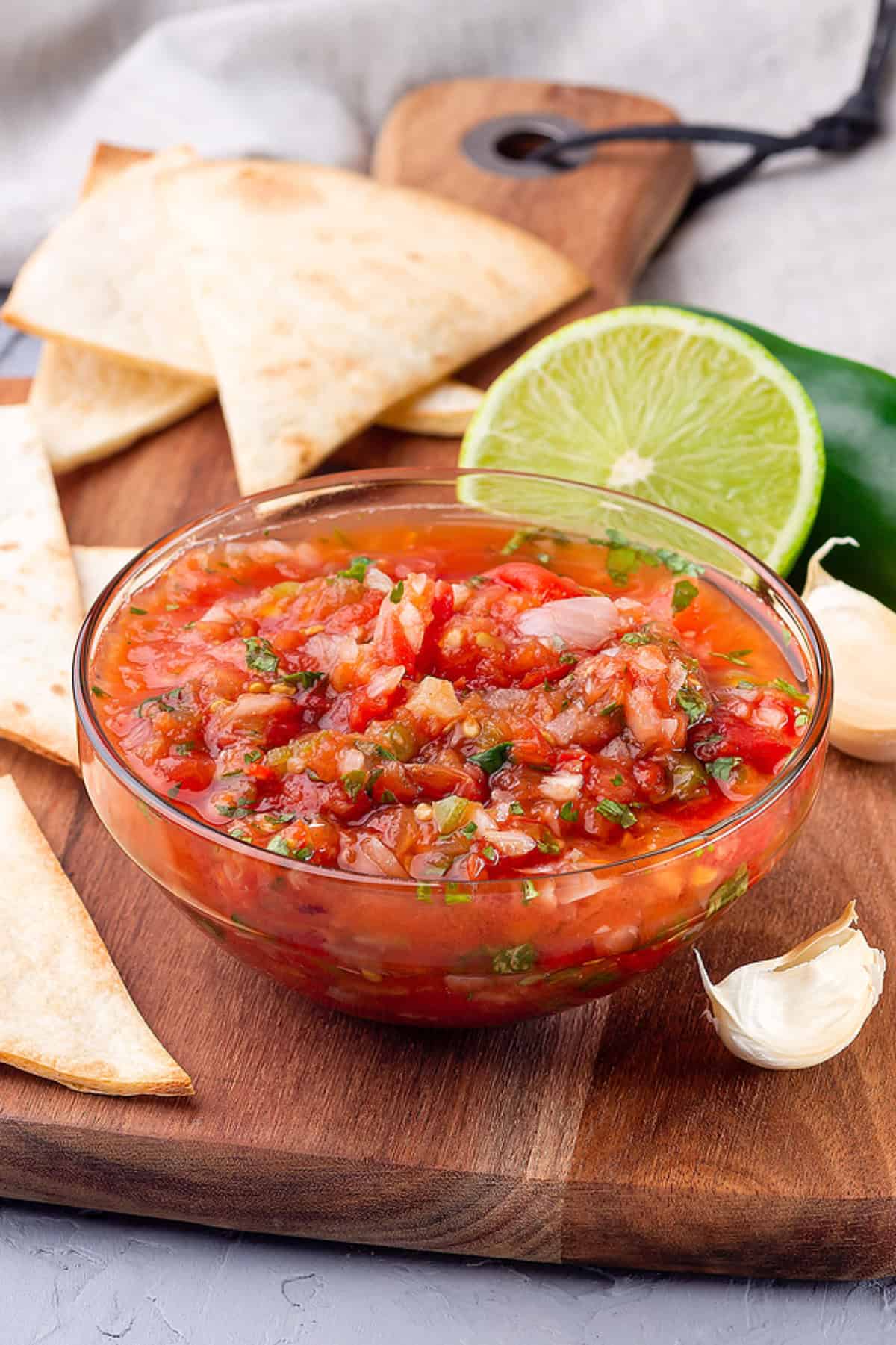Pico de gallo served on a wooden board with tortilla chips, lime, and garlic.