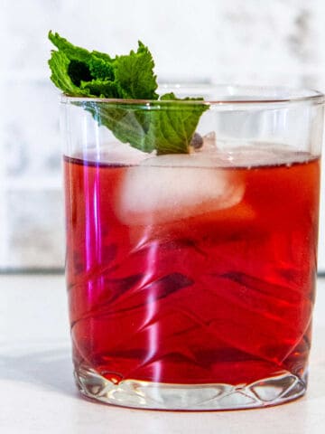 A glass of pomegranate compari spritz with ice cubes and a mint garnish sits on a white surface, with silver tinsel and lights in the background.