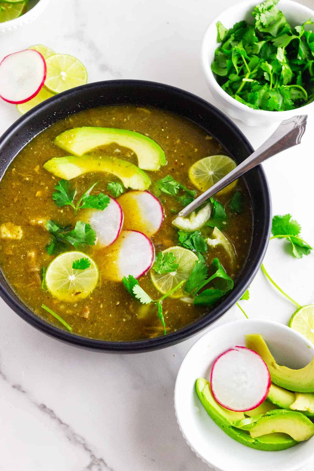 Pork pozole verde served with sliced avocado, radishes, lime wedges, and cilantro.