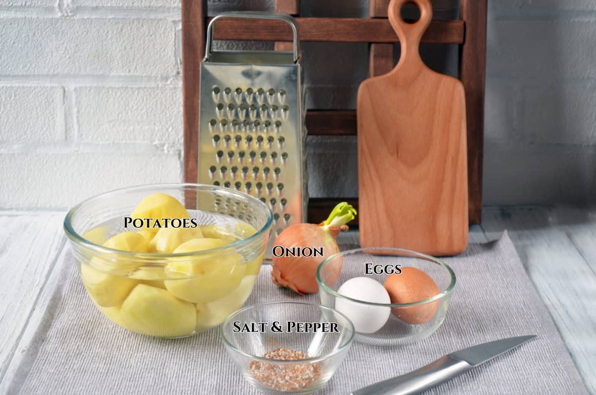 A glass bowl of peeled potatoes, a whole onion, two eggs, a small bowl of salt and pepper, a metal grater, cutting board, and knife arranged on a table with ingredient labels.