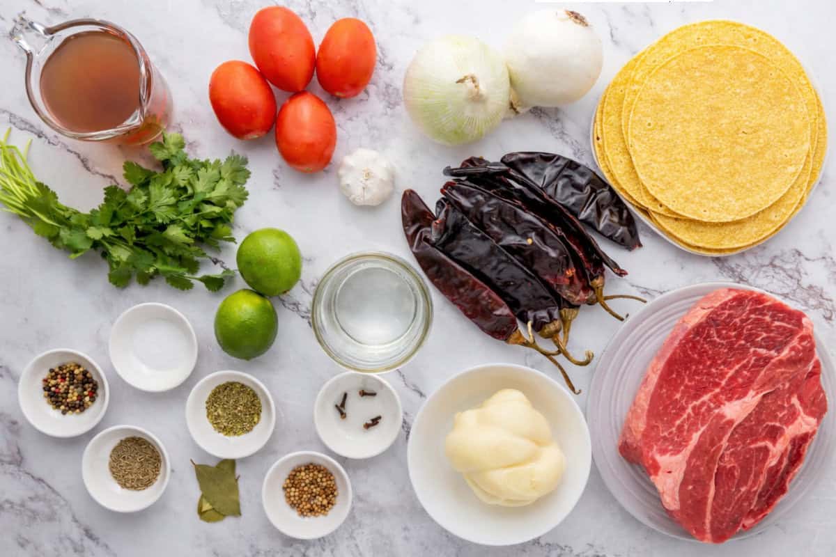 Assorted ingredients for birria laid out on a white surface, including raw beef, dried chilies, tomatoes, onions, tortillas, cilantro, limes, cheese, broth, and various spices in small bowls.