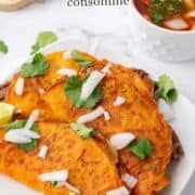Three cheesy, orange-colored quesabirria tacos topped with chopped onions and cilantro, served with lime wedges on a white plate. A bowl of rich consommé and a cutting board with more tacos appear in the background.