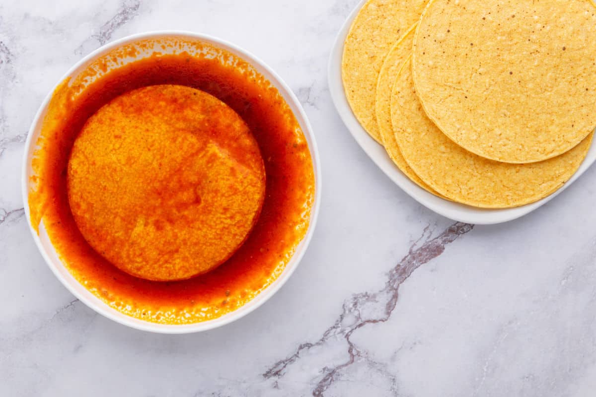 A white bowl filled with a corn tortilla soaked in red enchilada sauce sits on a marble surface next to a plate with several plain corn tortillas.