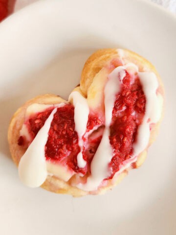 Heart-shaped crescent roll raspberry danish pastries are topped with red raspberry filling and drizzled with white icing, arranged on a light-colored surface.
