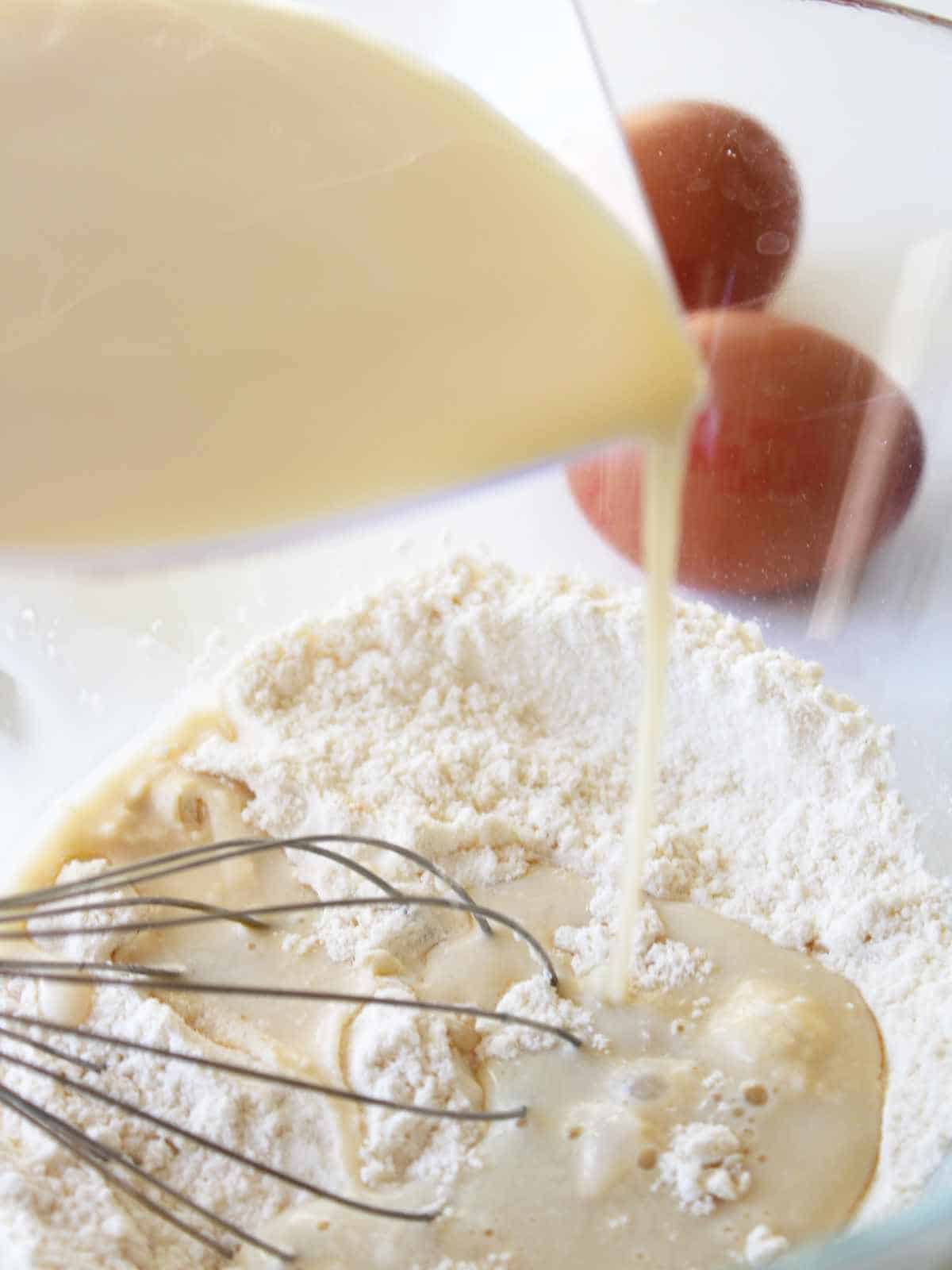 milk being poured into flour with eggs in the background.
