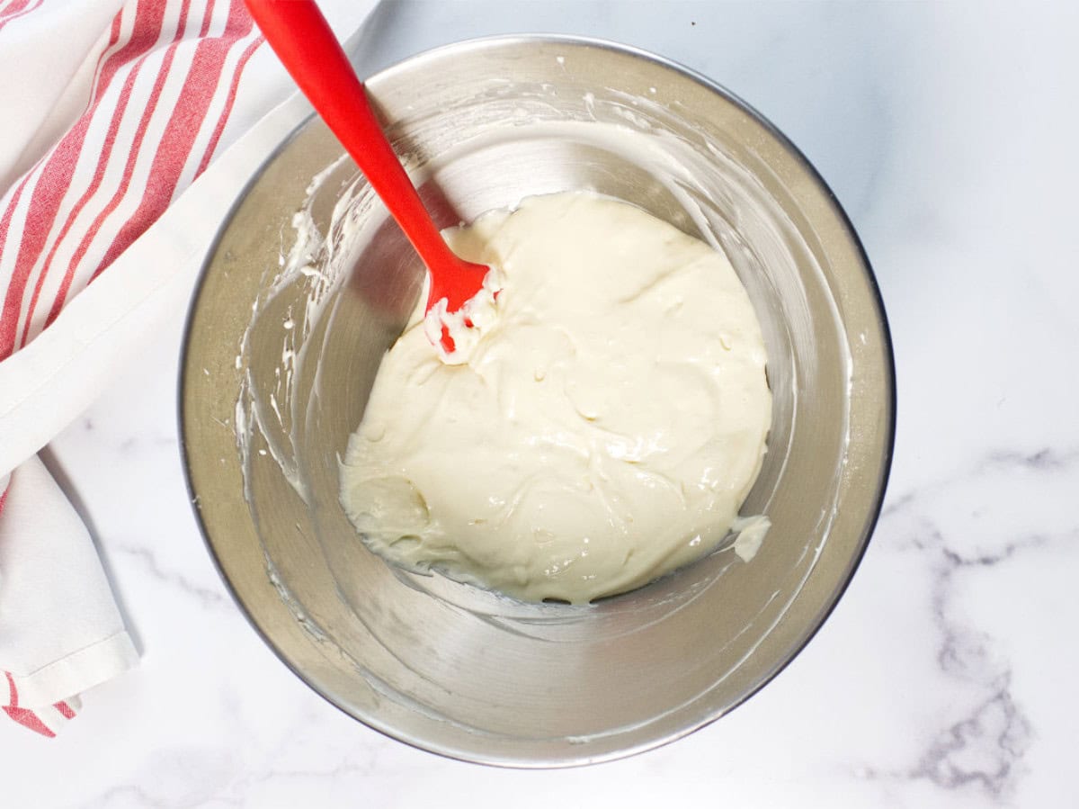 A metal mixing bowl with creamy Red Velvet Bundt Cake batter inside, being stirred with a red spatula. A white and red striped kitchen towel rests on the marble countertop nearby.