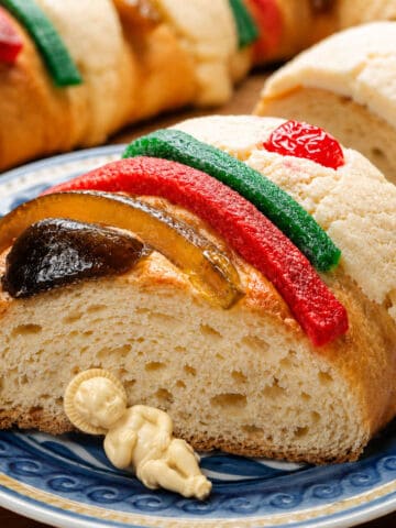 Close-up of Rosca de Reyes slice with candied fruit and cookie paste topping on a blue plate.