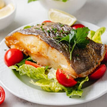 A cooked fish fillet with crispy skin served on a bed of lettuce and cherry tomato halves, garnished with fresh parsley and a lemon wedge on a white plate—a simple yet elegant choice for seafood recipes.