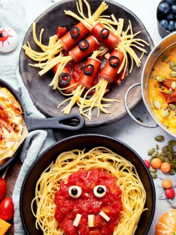 A variety of Halloween party treats, including pasta with monster faces, a pie with candy eyes, soup with seeds and shapes, plus scattered fruits and candies, all decorated to look spooky and fun.