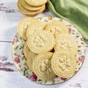 Round, scalloped-edge stamped shortbread cookies with a faint embossed pattern rest on a brown-patterned plate and a metal cooling rack, with a white and green cloth in the background.