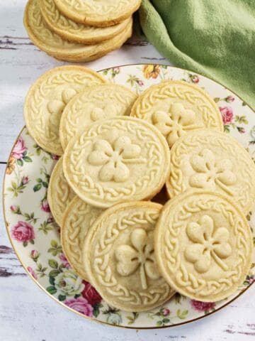 Round, scalloped-edge stamped shortbread cookies with a faint embossed pattern rest on a brown-patterned plate and a metal cooling rack, with a white and green cloth in the background.