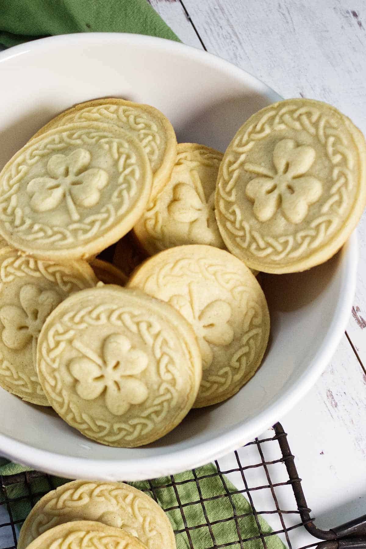 Six round stamped shortbread cookies on a decorative plate, resting on a wire cooling rack. A white kitchen towel with green stripes is in the background on a white wooden surface.
