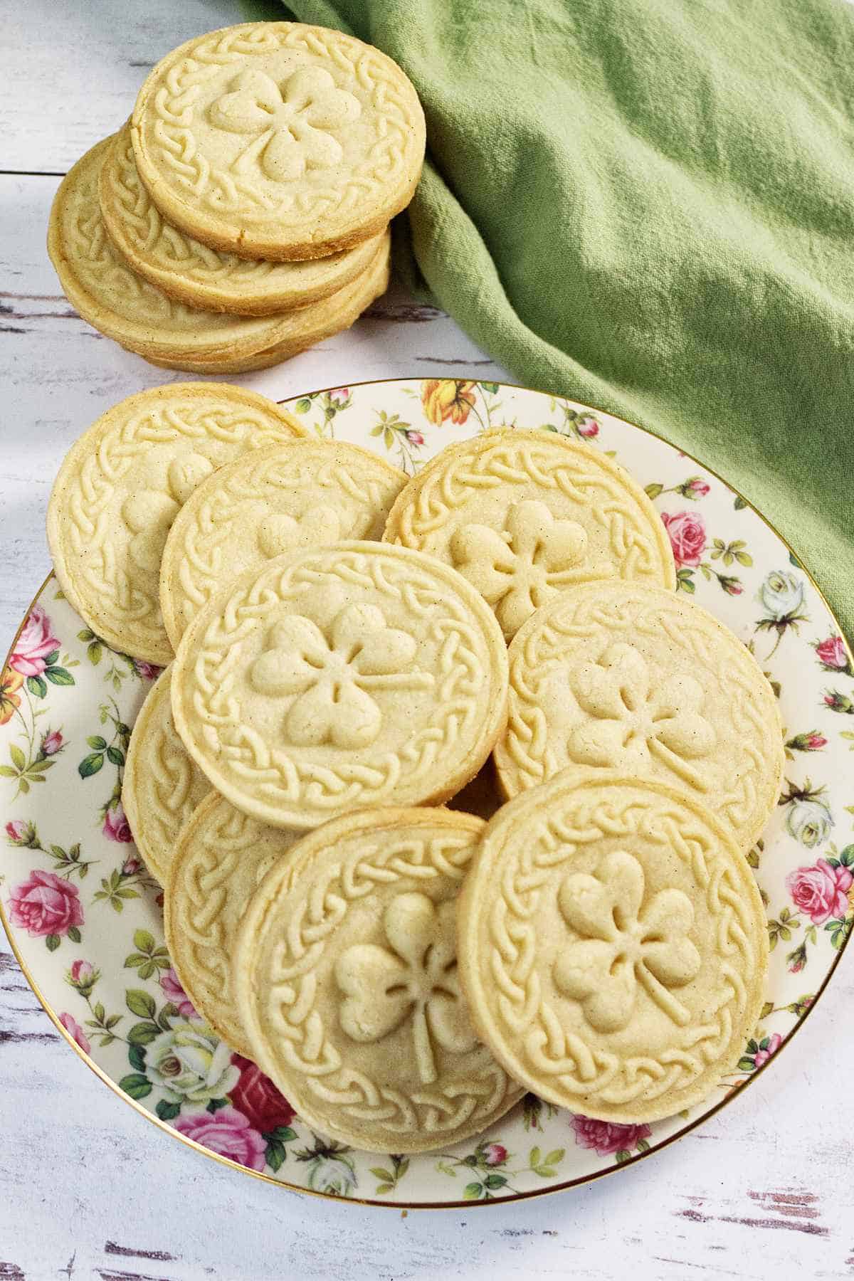 A plate of six round, golden stamped shortbread cookies with scalloped edges and embossed designs sits on a cooling rack, with a striped kitchen towel in the background.