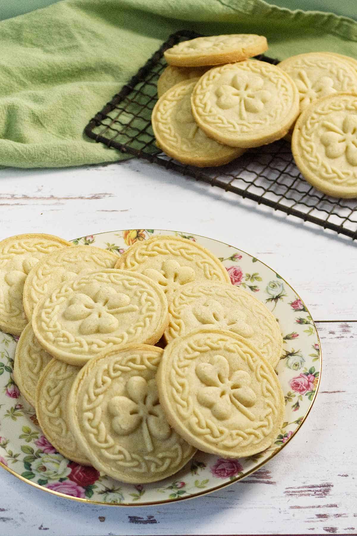 Six round, scalloped-edged stamped shortbread cookies rest on a metal cooling rack and a patterned plate, set on a white cloth with green stripes, atop a rustic white wooden surface.