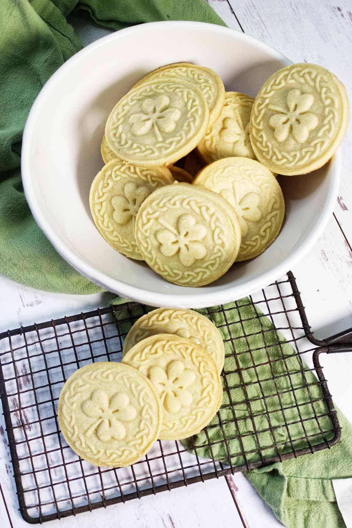 A stack of round, golden sugar cookies sits on a decorative plate, while stamped shortbread cookies with a faint embossed design rest on a cooling rack in the foreground.