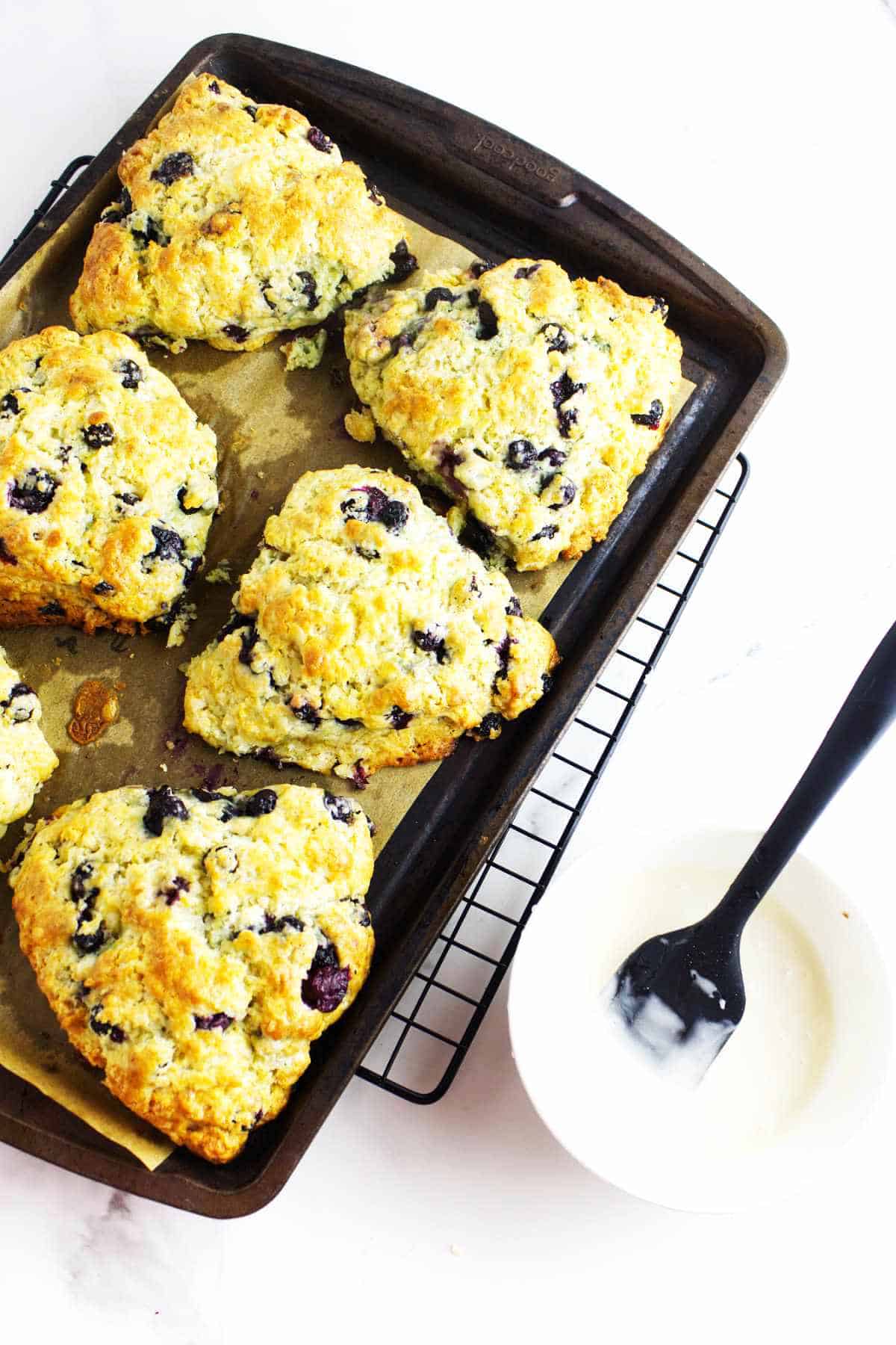 baking sheet of golden brown blueberry scones and a bowl of icing.