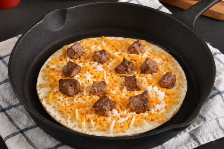 Steak and shredded cheese on a tortilla in a cast iron skillet.