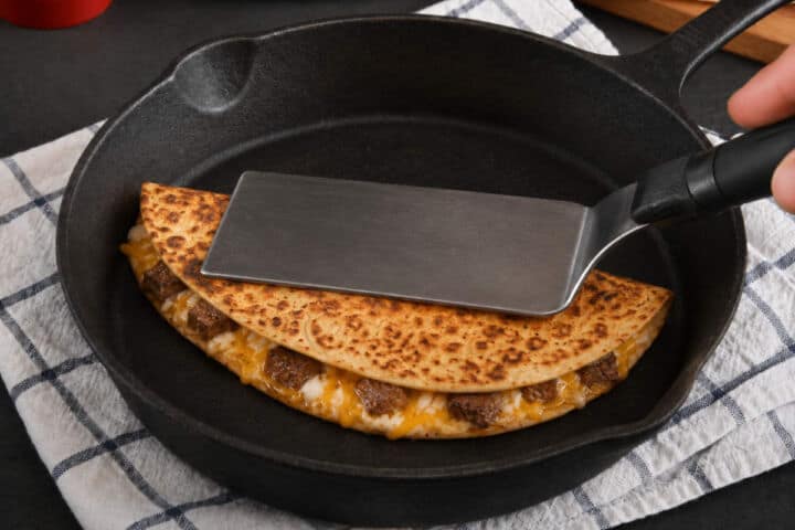 Spatula pressing a golden steak quesadilla in a cast iron skillet.