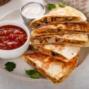 Three golden-brown steak quesadilla wedges filled with melted cheese and beef are garnished with chopped parsley on a white plate, with a dollop of sour cream on the side.