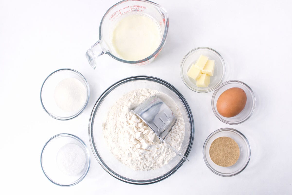 A top-down view of bowls and a measuring cup containing labeled ingredients for fluffy dinner rolls—flour, milk, butter, yeast, salt, and sugar—arranged neatly on a white surface.