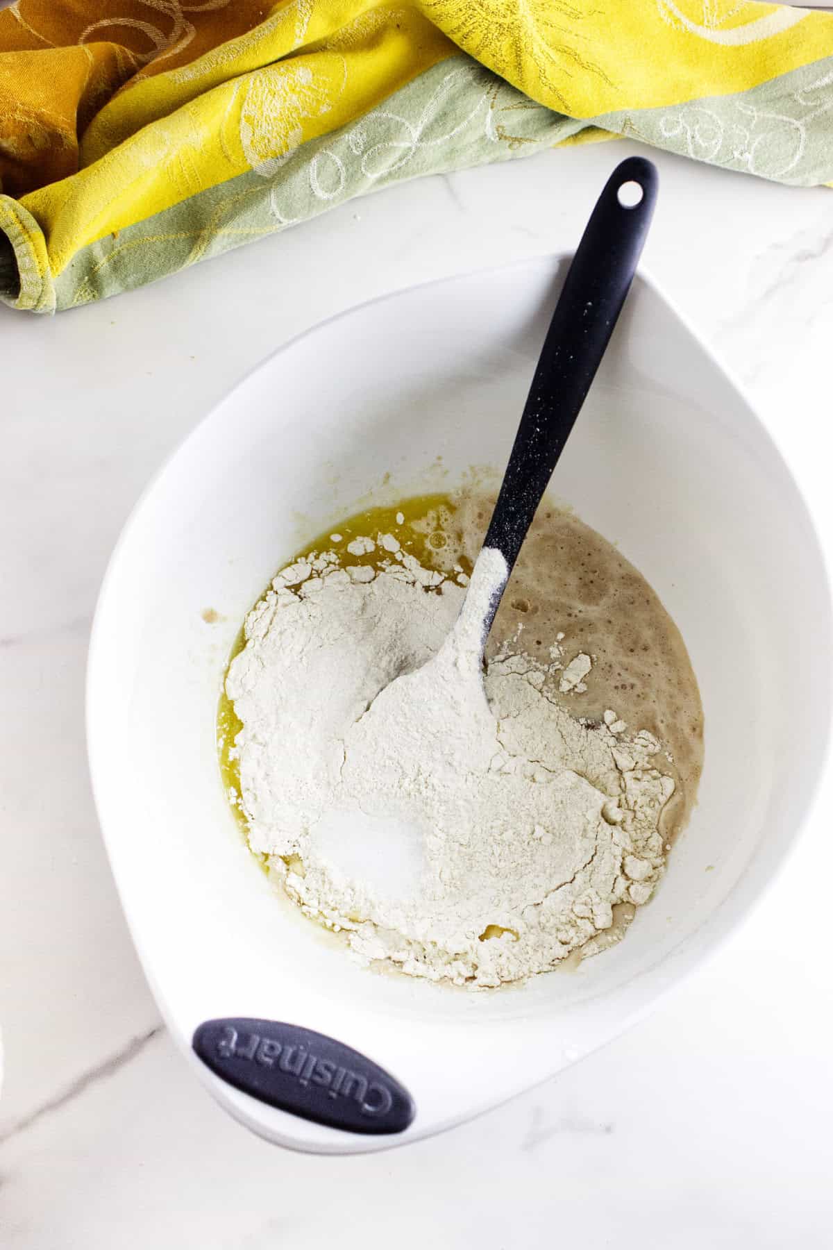 A white mixing bowl with flour, yeast mixture, and melted butter inside-perfect beginnings for fluffy dinner rolls. A black spoon rests in the bowl, while a yellow and green cloth sits atop the white countertop.