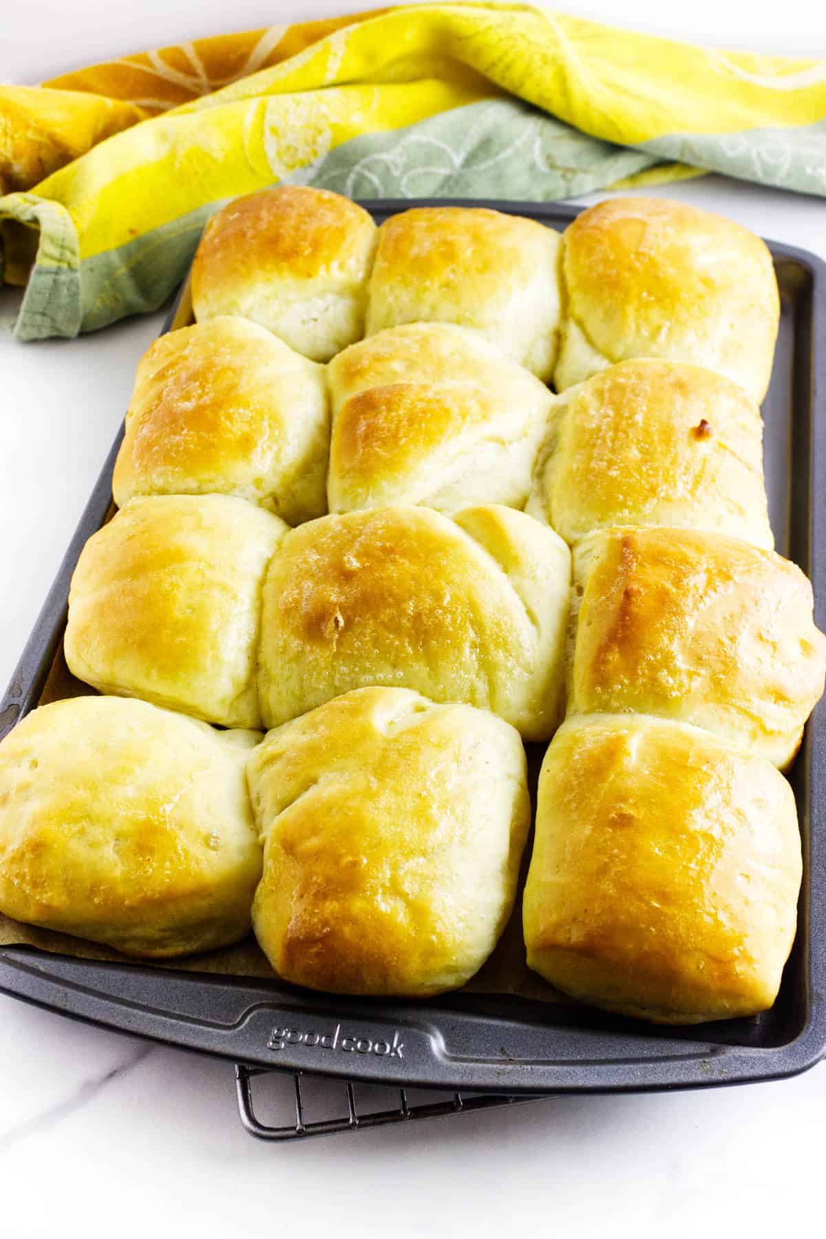 A baking tray filled with twelve fluffy dinner rolls, perfectly golden-brown and freshly baked, sits in front of a yellow and green kitchen towel.