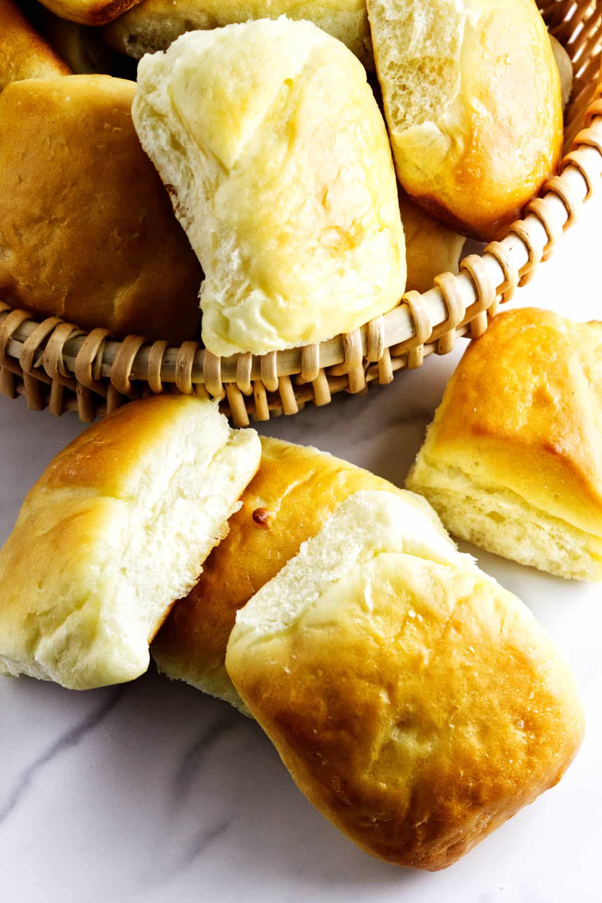 A basket filled with golden, fluffy dinner rolls, with a few fluffy dinner rolls placed on a white marble surface beside the basket. One roll in the foreground is split open, showing its irresistibly soft interior.