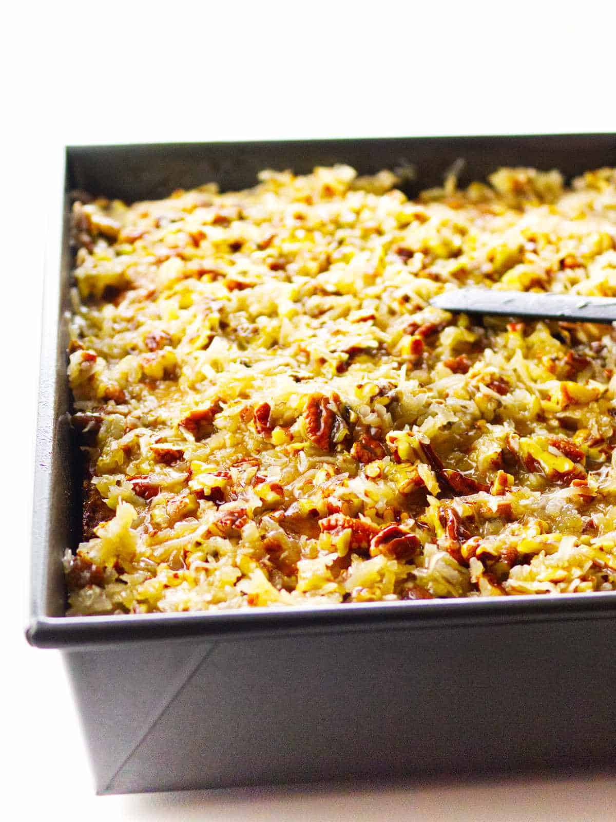 A close-up of a rectangular baking pan filled with golden-brown Texas Tornado Cake, topped with shredded coconut and chopped pecans. A knife rests on top of the dessert.