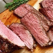 Sliced cooked beef roast with a peppery crust, arranged on a wooden cutting board. The meat is juicy with a pink center and seasoned outer layer.