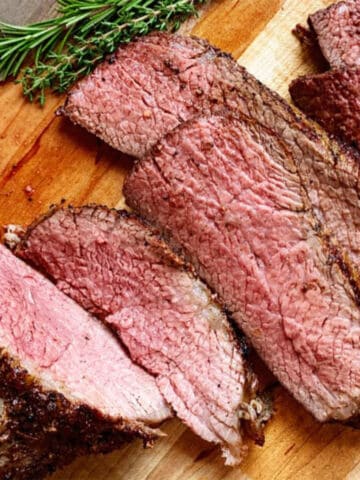 Sliced cooked beef roast with a peppery crust, arranged on a wooden cutting board. The meat is juicy with a pink center and seasoned outer layer.