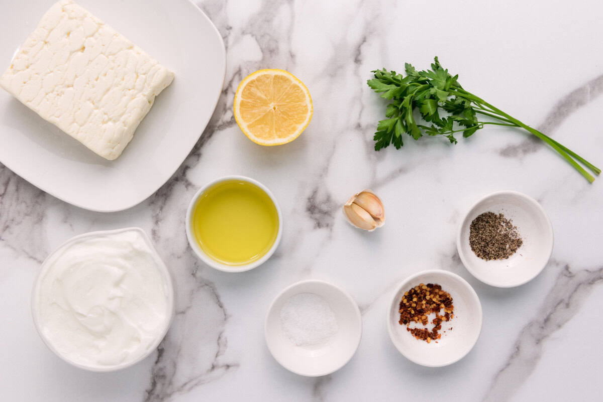 A marble surface with whipped feta cheese dip, half a lemon, parsley, a bowl of olive oil, a garlic clove, Greek yogurt, and small bowls of salt, crushed red pepper, and black pepper.