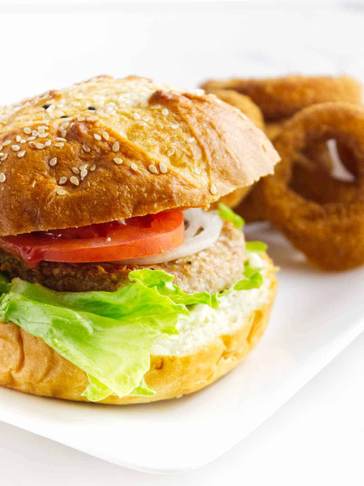 turkey burger on a bun with slices of tomato, onion, and lettuce with onion rings nearby.