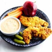 healthy child's lunch plate with apple, carrot sticks, pickles, and air fryer chicken tenders with dip.