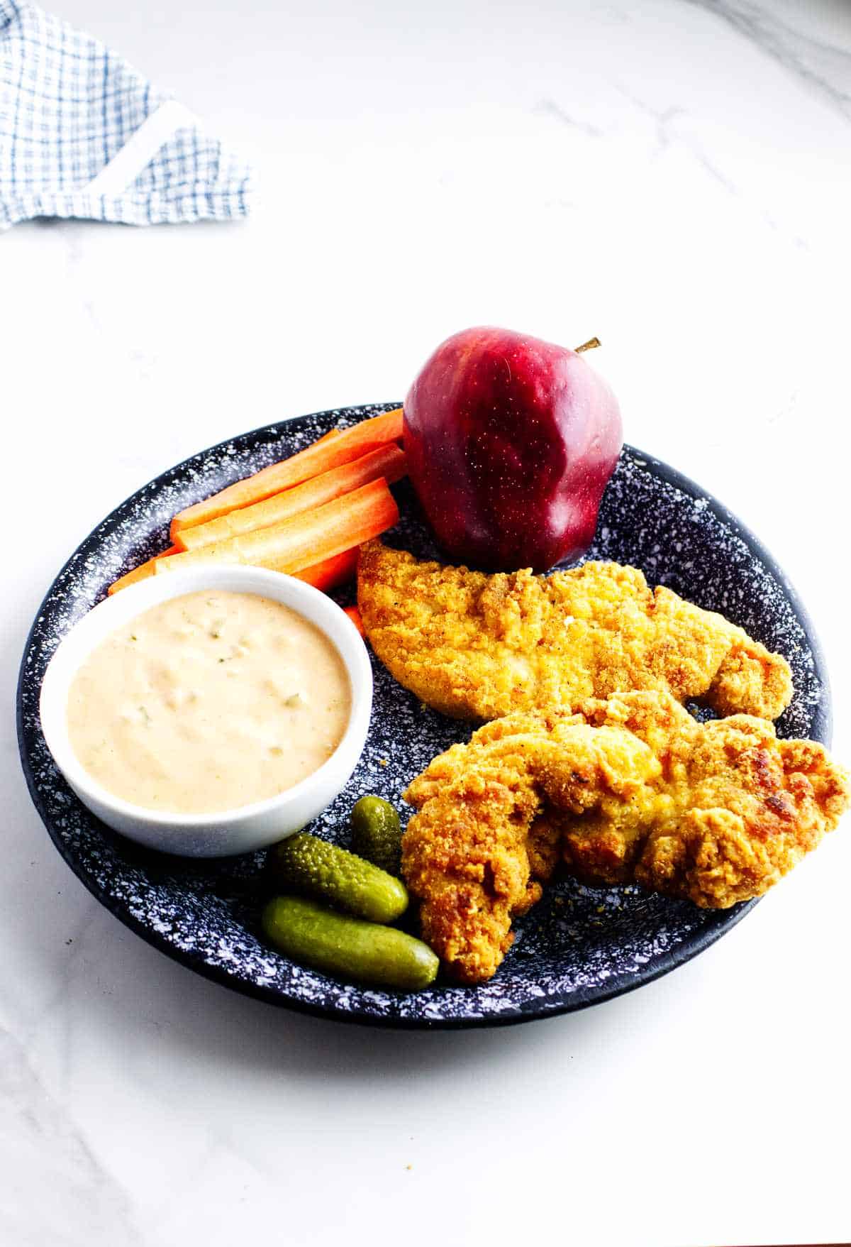 healthy child's lunch plate with apple, carrot sticks, pickles, and air fryer frozen chicken tenders with dip.