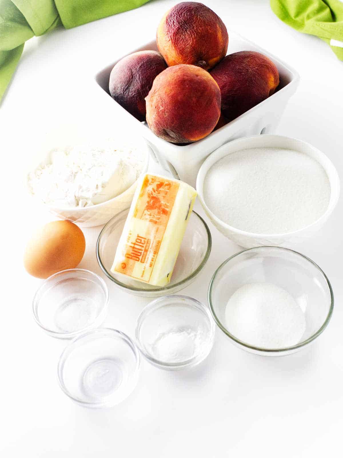 basket of peaches, cube of butter, an egg, and flour, sugar, and milk on a white background.
