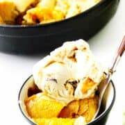 peach cobbler in a bowl with ice cream on top, with cast iron peach cobbler in background.