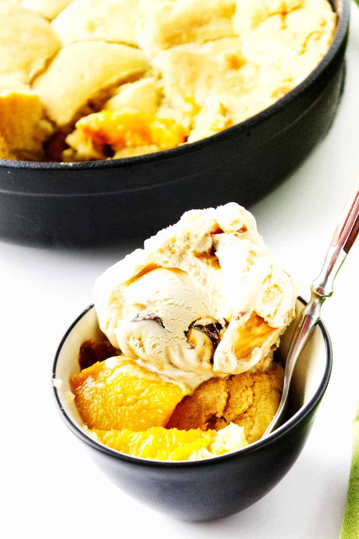 peach cobbler in a bowl with ice cream on top, with cast iron peach cobbler in background.